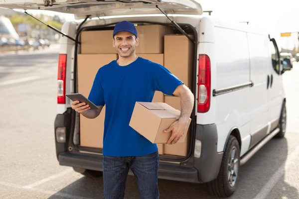 Young European Man Courier In Uniform Using Touchpad And Holding Parcel Box, Standing Outdoors Near Van